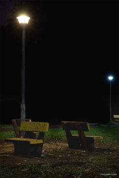 two park benches sitting under a street light at night with the lights turned on in the background