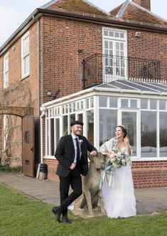 a bride and groom walking with their dog in front of a large brick building on the lawn