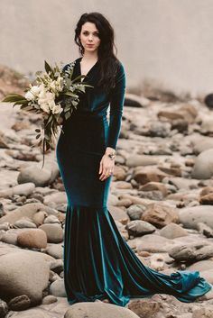 a woman in a long green dress standing on rocks holding a bouquet and looking off into the distance