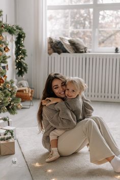 a woman holding a baby in her arms while sitting on the floor next to a christmas tree