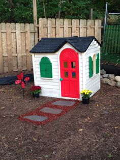 a small white house with red door and green shutters on the side, in front of a wooden fence