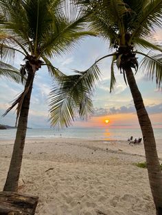 the sun is setting behind two palm trees on the beach with people in the distance