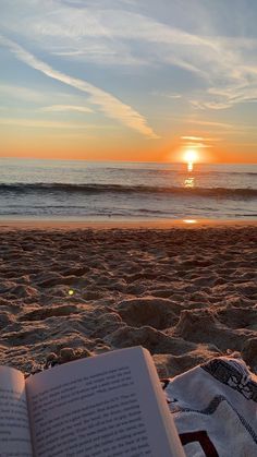 an open book sitting on top of a sandy beach next to the ocean at sunset