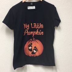 This Is A Brand New T-Shirt That Was Never Worn. A Great Way To Announce Your Pregnancy Or To Wear At The Pumpkin Patch This Fall. Plain Black T-Shirt With My Little Pumpkin On The Front And Cute Little Feet Inside The Pumpkin. Just Precious. Cute Black T-shirt With Screen Print, Cute Pre-shrunk Black T-shirt, Cute Black Pre-shrunk T-shirt, Black Pre-shrunk T-shirt For Fall, Black Family Matching Tops For Fall, Family Matching Black Tops For Fall, Black Logo Print T-shirt For Fall, Cute Black T-shirt With Text Print, Cute Orange T-shirt With Letter Print