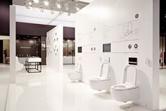 three toilets are on display in a room with white walls and flooring, along with tables and chairs