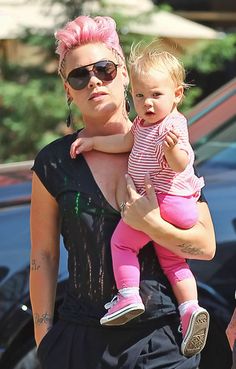 a woman with pink hair holding a baby in her arms and looking at the camera