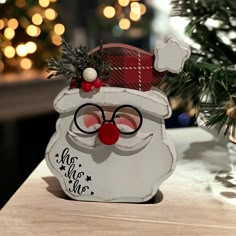 a wooden santa clause with glasses and a red nose on a table next to a christmas tree