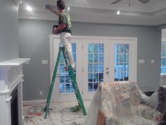 a man standing on a ladder painting the ceiling in a room with gray walls and white trim