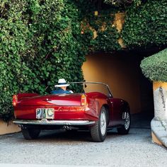 an old red car parked in front of a green hedge covered building with a white hat on top