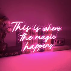 this is where the magic happens neon sign on a shelf next to a vase with flowers