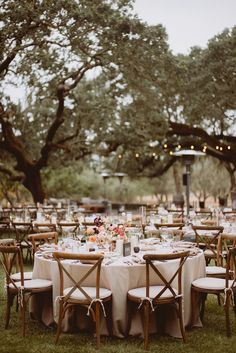 an outdoor table set up with white linens and wooden chairs, surrounded by trees