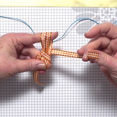 two hands are tying a piece of fabric with orange and white checkered ribbon on a graph paper background