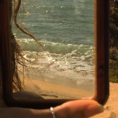 a person's hand holding up a mirror to take a photo of the ocean