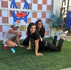 three women sitting on the grass in front of a building and posing for a photo