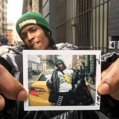 a man holding up a polaroid photo in front of a car on the street