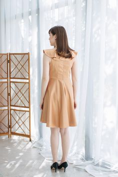 a woman standing in front of a window with her back to the camera, wearing a tan dress