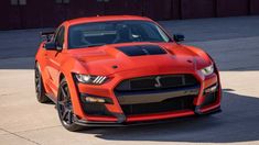 an orange sports car parked in front of a red building with black stripes on it's hood