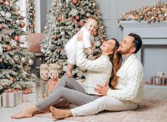 a man and woman sitting on the floor with their baby in front of christmas trees