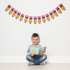 a young boy sitting on the floor reading a book with pencils hanging above him