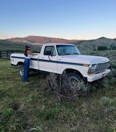 a woman standing next to a white pickup truck in a field with mountains in the background