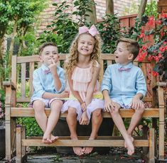 three children sitting on a wooden bench in front of some bushes and flowers with their mouths open