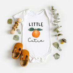 a baby bodysuit that says little cutie with an orange on it next to shoes and plants