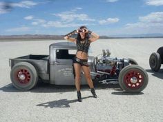 a woman standing next to an old truck in the middle of a barren area with another car behind her