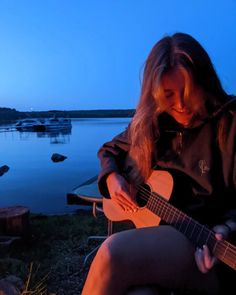 a woman is playing an acoustic guitar by the water at night with boats in the background