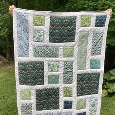 a person holding up a quilt in front of some trees and bushes with green grass