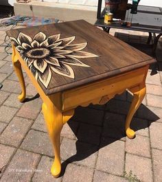 a wooden table with a sunflower painted on it's top sitting on a brick sidewalk