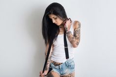 a woman with tattoos and suspenders is holding her hand on her shoulder while standing against a white wall