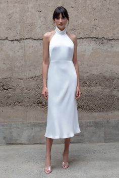 a woman standing in front of a stone wall wearing a white dress with high neck