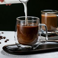 someone pouring milk into a glass mug filled with coffee