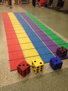 three dices are on the ground in front of a rainbow colored mat with people standing around