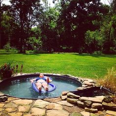 a man laying in an outdoor hot tub surrounded by rocks and grass with trees in the background