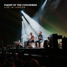 two men on stage playing instruments in front of an audience at a concert with bright lights