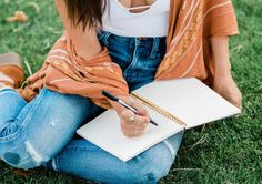 a woman sitting on the grass holding a notebook and pen in her hand while writing