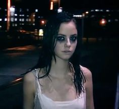 a woman with black makeup and dark eyes standing in the street at night, wearing a white tank top