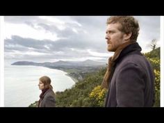 two people standing on top of a hill next to the ocean and hills with yellow flowers