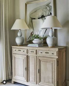 two lamps sitting on top of a wooden dresser next to a painting and lamp shades