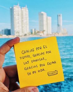 a person holding up a yellow post it note in front of the ocean with buildings in the background