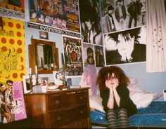 a woman sitting on top of a bed next to a dresser and wall covered in posters