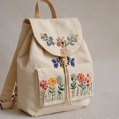 a white backpack with embroidered flowers on the front and side pockets, hanging from a leather strap