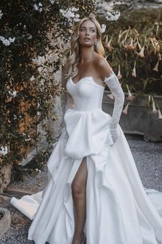 a woman wearing a white wedding dress with long sleeves and thigh high slits standing in front of a bush