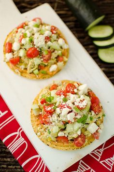 two small pizzas sitting on top of a cutting board next to cucumbers