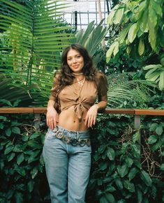 a woman standing in front of green plants wearing jeans and a shirt with a tie around her waist