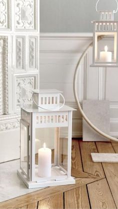 a white lantern with two lit candles in it on a wooden floor next to a mirror