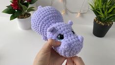 a hand holding a purple crocheted hippo stuffed animal next to potted plants