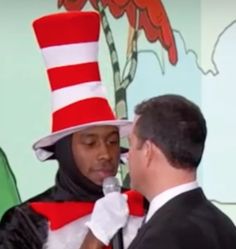 a man wearing a cat in the hat talking into a microphone while standing next to another man