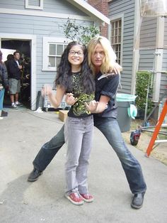 two women are posing for the camera in front of a house with people standing around